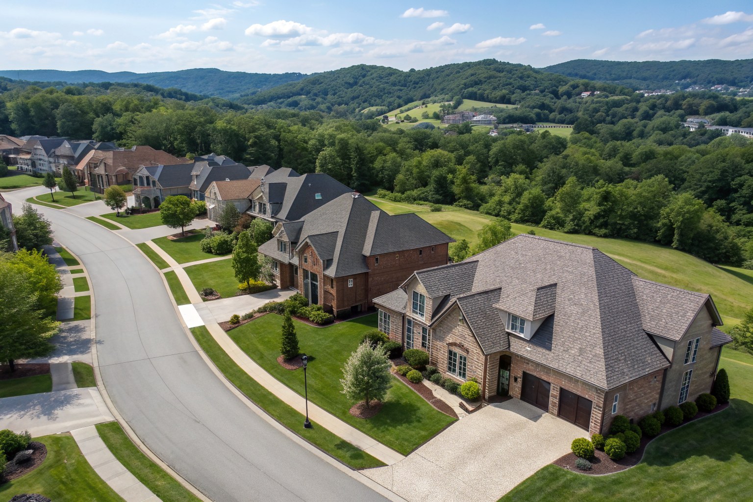 Aerial view of large brick homes with gray and brown shingle roofs along a curving street among rolling hills